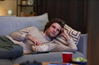 © Seventyfour - Portrait of young man watching Tv at home and eating popcorn while lying on couch, copy space