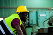 © KANGWANS - A black man African American worker wearing glasses in a yellow hard hat works machinery in an industrial factory.