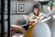© nenetus - Pretty young woman reading a book while sitting on sofa at home.