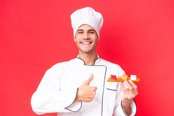 Wall Mural - Young caucasian chef man holding sashimi isolated on red background giving a thumbs up gesture