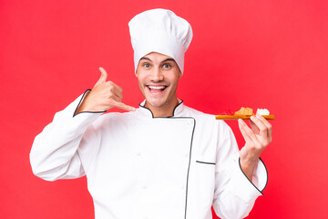 Wall Mural - Young caucasian chef man holding sashimi isolated on red background making phone gesture. Call me back sign