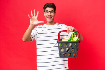 Wall Mural - Young caucasian man man holding a shopping basket full of food isolated on red background counting five with fingers