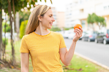 Wall Mural - Young blonde woman holding an orange at outdoors with happy expression