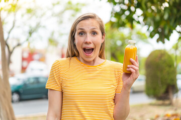 Young blonde woman holding an orange juice at outdoors with surprise and shocked facial expression