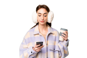 Wall Mural - Young caucasian woman wearing winter muffs over isolated chroma key background holding coffee to take away and a mobile