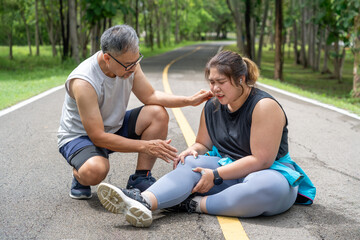 Wall Mural - Overweight young woman with injured knee sitting on the running track at local park with both hands grabbing on her trouble knee while a middle age man kneeling beside her