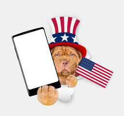 Poster - Serious Mastiff puppy wearing like Uncle Sam looking through a hole in paper and showing big smartphone with white blank screen. isolated on white background