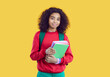 Smiling cute dark skinned preteen schoolgirl with backpack and notebooks on yellow background. Portrait of pretty ethnic curly teen girl who is ready to go to school. Concept of school education.