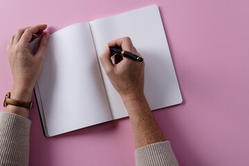 Poster - Hands of caucasian woman holding pen and writing in notebook with copy space on pink background