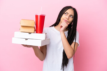 Poster - Teenager girl with braids holding pizzas and burgers over isolated pink background and looking up