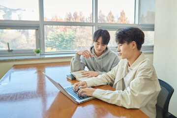 A male student sits at a desk in a university library in South Korea, Asia, looking at a laptop computer with bookshelves of books in the background.