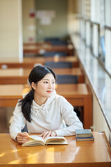 Asian Korean University Library Young female college student model sitting at a desk by a window with light coming in, reading a book, looking out the window