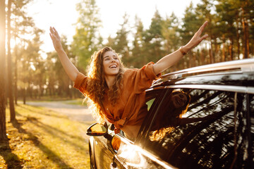 Beautiful traveler woman is resting and enjoying the sunset in the car. Happy woman on a summer trip, travels leaning out of the car window. Active lifestyle, tourism. Travel concept.