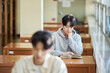 © TEAM PRE-LIGHT - Three college students, both male and female, are sitting or standing at a desk in a library at a South Korean university in Asia, looking at laptops and books while having a conversation.