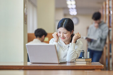 Young Asian Korean female model in library looking at laptop or book, lecture, assignment, discussion, male model in background, bookshelf in background