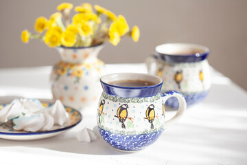 Ceramic mugs with tea on the kitchen table. There is a bouquet of yellow flowers in the background. Selective focus. 