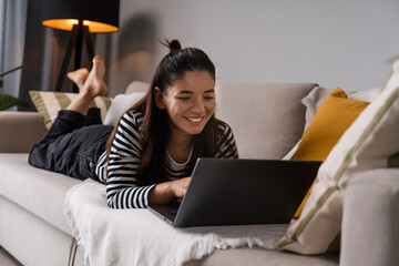 Wall Mural - Relaxed study. Attractive student lying on sofa bed cheerfully working on laptop