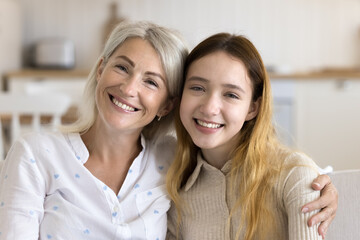 Wall Mural - Cheerful attractive blonde grandma and pretty teenage kid home head shot portrait. Positive grandmother and child enjoying family meeting, sitting close, looking at camera, hugging