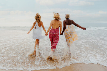 view from behind of multiracial group of young women on the shore of the beach with their hands together going into the sea. friendship and summer concept