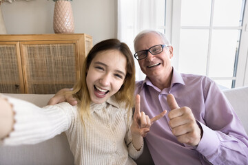 Wall Mural - Self portrait of happy excited teenager granddaughter kid and cheerful older grandpa showing hand symbols, gestures at camera, smiling, laughing, having fun, taking selfie together at home