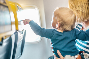 Mom and child flying by plane. Mother holding and playing with her infant baby boy child in her lap during economy comercial flight. Concept photo of air travel with baby. Real people