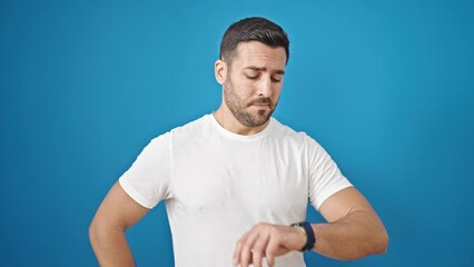 Poster - Young hispanic man looking watch with relaxed expression over isolated blue background