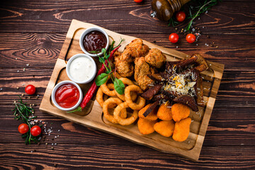 Wall Mural - Snacks breaded squid rings, chicken nuggets, rye bread croutons with cheese, chicken legs and sauces on a wooden board.