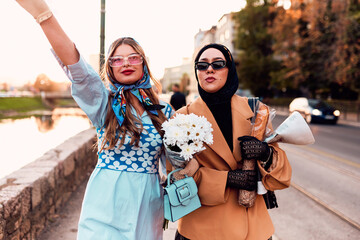 Couple woman one wearing a hijab and a modern yet traditional dress, and the other in a blue dress and scarf, walking together through the city at sunset. One carries a bouquet and bread, while the