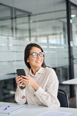 Young professional Asian business woman worker holding cellphone using mobile looking away thinking on new cell phone tech solution feeling happy sitting at desk in office working, vertical.