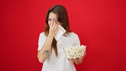 Wall Mural - Young beautiful hispanic woman watching drama movie eating popcorn crying over isolated red background