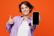 © ViDi Studio - Young woman she wears purple shirt white t-shirt casual clothes hold in hand use close up mobile cell phone with blank screen workspace area show thumb up isolated on plain orange background studio.
