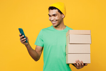 Wall Mural - Young man of African American ethnicity wears casual clothes green t-shirt hat hold stack cardboard blank boxes use mobile phone isolated on plain yellow background studio portrait. Lifestyle concept.