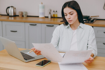 Young businesswoman housewife latin woman wears casual clothes striped shirt work with laptop pc computer bookkeeping documents sit at table in kitchen at home alone. Lifestyle cooking food concept.