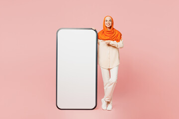 Full body young arabian muslim woman wear orange abaya hijab point index finger on big huge blank screen mobile cell phone with area isolated on plain pink background Uae middle eastern islam concept