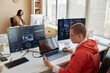 © Seventyfour - Side view of young female cyber security engineer using smartphone while sitting by workplace in front of three computers with data on screens