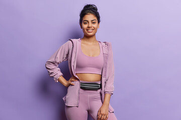 Horizontal shot of pleasant looking sporty young woman with athletic body keeps hand on waist smiles gladfully dressed in sportswear isolated over purple background. Sporty lifestyle concept