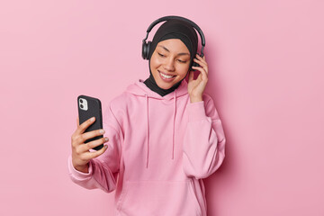 Wall Mural - Photo of pleasant looking young Muslim woman wearing headphones for listening to music via mobile phone makes video call wears traditional hijab and sweatshirt isolated over pink backgroundd