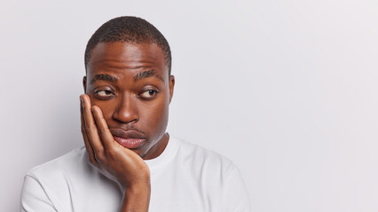 Photo of displeased sad dark skinned man keeps hand on cheek looks thoughtfully aside has dejected frustrated expression wears casual t shirt isolated over white background copy space for your text