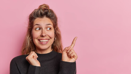 Wall Mural - Horizontal shot of positive woman with curious expression toothy smile on face points finger on blank space rejoices positive news shows copy space for advertisement isolated over pink background