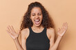 Overjoyed Brazilian woman with curly hair raising her palms and exclaiming loudly in amazement keeps mouth widely opened wears black t shirt isolated over brown background. Human reactions concept