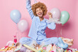 Cheerful joyful curly haired woman dressed in pajamas enjoys domestic party celebrates anniversary surrounded by colorful balloons stands near table filled with delectable desserts dances carefree