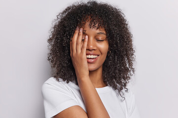 Wall Mural - Curly haired woman radiates positivity as she covers half her face with her palm eyes closed in blissful contentment dressed in casual tshirt isolated on white background. People and happiness concept