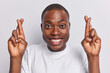 Studio shot of dark skinned short haired man crossing his fingers in gesture of hope and anticipation exudes cheerful and positive energy smiles toothily dressed casually isolated on white background