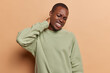 Displeased dark skinned man suffers from neck ache clenches teeth keeps eyes closed feels tension wers casual green jumper isolated over brown background. People and health problems concept.