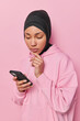 People and religion concept. Serious Muslim woman holds chin concentrated at smartphone screen with attentive gaze reads news online wears black hijab and hoodie isolated over pink background