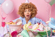 Startled young woman with curly hair wears cone hat and pajamas poses near festive table with desserts celebrated birthday isolated over pink background. People celebration and partying concept