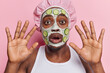 Isolated portrait of scared frightened man applying cucumber slices and clay mask doing morning spa procedure keeps palms raised up wears bath hat and white t shirt isolated over pink background