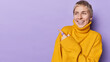 Happy short haired woman smiles gladfully points index finger at copy space advertises something dressed in casual warm yellow jumper isolated over purple background. People and promotion concept
