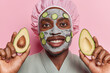 Pleased African man delicately applies cucumber adorned beauty masks on his face holds avocado halves smiles toothily wears bath hat and dressing gown stands against pink background. Beauty concept