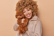 Cheerful curly female pet owner exudes love and warmth as she holds poodle puppy in her hands dressed in cozy winter coat showcasing tbond between humans and their furry companions poses indoor